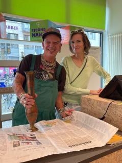 Der Berliner Friseur Frank Schäfer (links), hat sich am „Mach etwas Nettes“-Tag am 5. Oktober 2023 im Oxfam Shop Prenzlauer Berg, Berlin, engagiert. Für eine Schicht hat er Ehrenamtliche wie Martina Günther (rechts) unterstützt. Ein Mann und eine Frau stehen lächelnd hinter der Kasse eines Oxfam Shops. Der Mann hält eine Vase in der Hand.