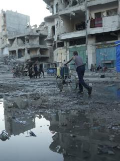 Ein Mann schiebt einen Karren mit Kanistern durch die Trümmer in Gaza-Stadt. Der Weg ist überschwemmt mit Abwasser. Ein Mann schiebt einen Karren mit Kanistern durch die Trümmer in Gaza-Stadt. Der Weg ist überschwemmt mit Abwasser. Im Hintergrund Ruinen und überall Schutt.