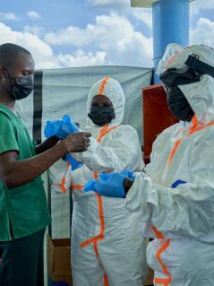 Vorbereitung eines Hygieneteams vor dem Betreten einer Ebola-Station: Joseph Kandule hilft dem Team in Schutzanzügen beim Anziehen der Schutzhandschuhe. Ein Mann in grünem T-Shirt und mit Mund-Nasen-Schutz hilft zwei Personen in weißen Schutzanzügen und mit Mund-Nasen-Schutz in ihre blauen Schutzhandschuhe.