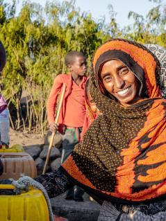 Kommt dank Wasserquelle besser durch den Alltag: Seadiya Mohamed Seadiya Mohamed lächelt in die Kamera, während sie Wasser aus einem laufenden Hahn in ihren Kanister schöpft. Im Hintergrund stehen zwei Kinder.