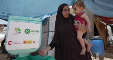 Duaa Abu Sabha benutzt eine Handwaschstation, die Oxfam für Geflüchtete im Gebiet Al-Mawasi im Gouvernement Khan Yunis im Gazastreifen aufgestellt hat. Duaa Abu Sabha wäscht sich die Hände an einer Handwaschstation, die in einem Camp für Geflüchtete steht. Auf dem Arm trägt sie ein Kleinkind.