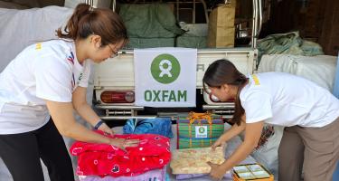 Zwei weiblich gelesene Personen in Oxfam-Shirts packen Kleidungsstücke und Handtücher zusammen.