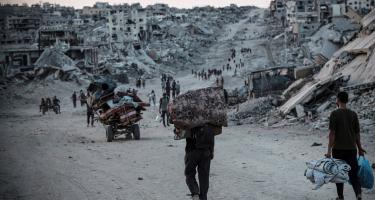 Menschen kehren zurück nach Jabalia in Nord-Gaza. Hier erwartet sie eine katastrophale Zerstörung. Menschen mit Taschen und Beuteln und Karren gehen einen Weg entlang. Rechts und links sind bis zum Horizont Trümmer und zerstörte Häuser zu sehen.