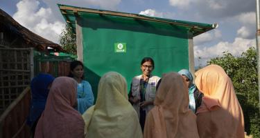 Sani Tweaks: Eine Frauengruppe, die mehrere sanitären Einrichtungen im Geflüchtetencamp Cox's Bazar (Bangladesch) mitgestaltet hat, spricht mit Iffat Tahmid Fatema über ihre Erfahrungen in den ersten Wochen der Nutzung der Latrine und der Dusche. Eine Frauengruppe steht vor einer Latrine im Kreis und spricht.